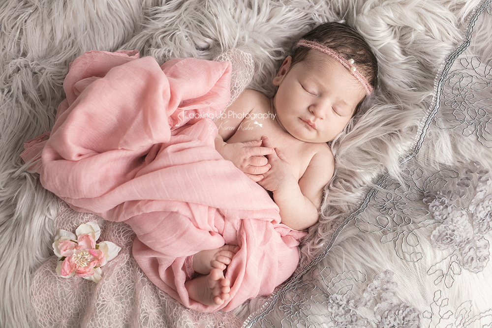 Sleeping newborn adorned in pink fabric and pearl headband surrounded by gray lace and cozy fur with gentle hands posed for baby girl portrait session