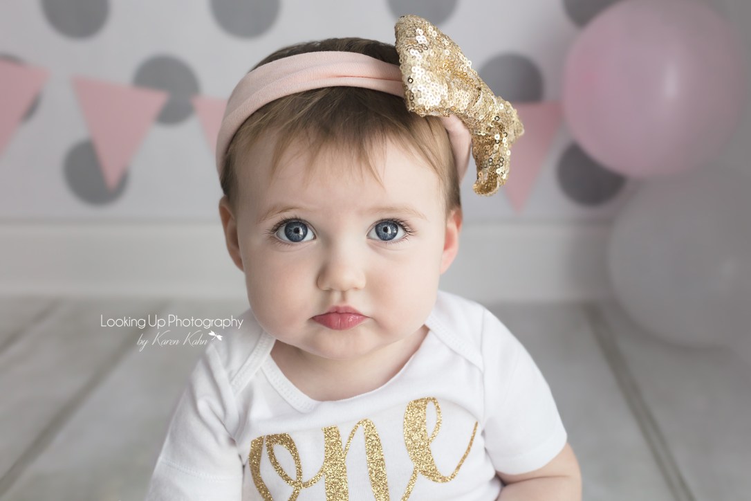 Pouty cuteness in gold and pink themes themed cake smash session for one year old milestone baby girl looking adorable in onesie with gray polka dots for 12 month portrait