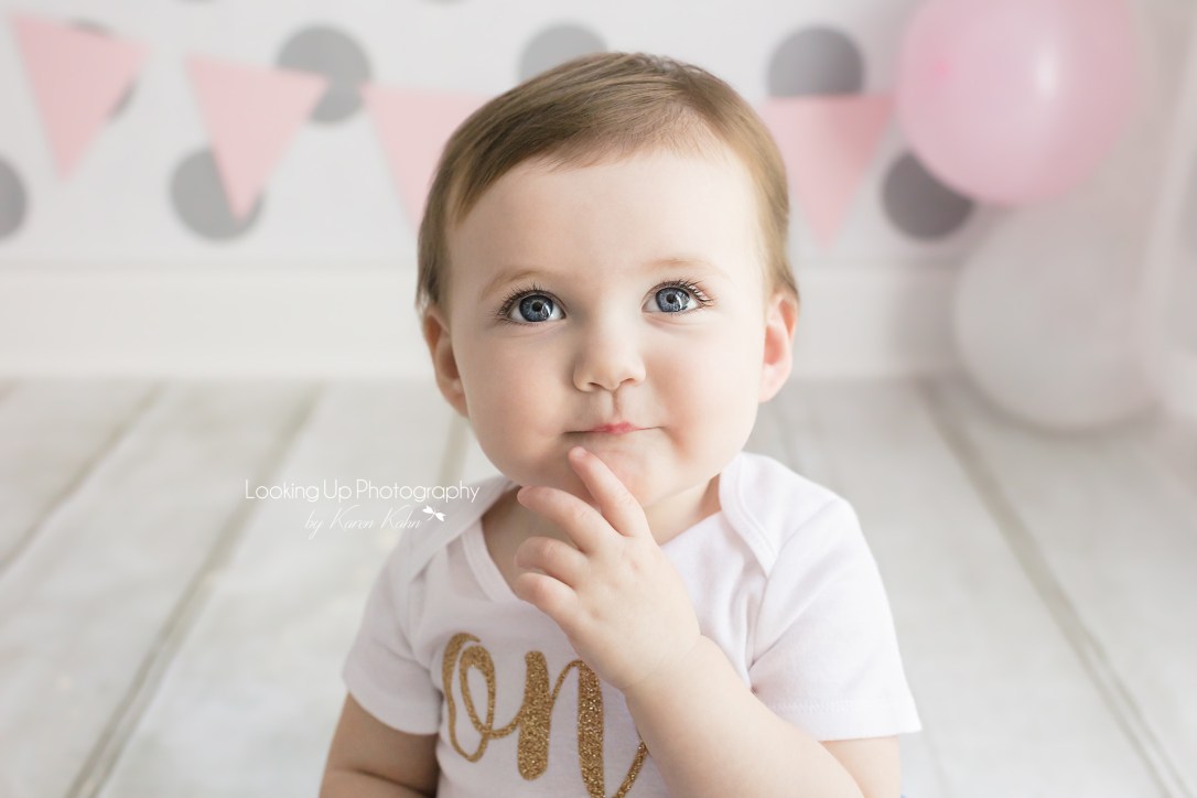 Thoughtful pose and big blue eyes in gold, pink and white cake smash session for one year old milestone baby girl looking sweet with gray polka dots for 12 month portrait