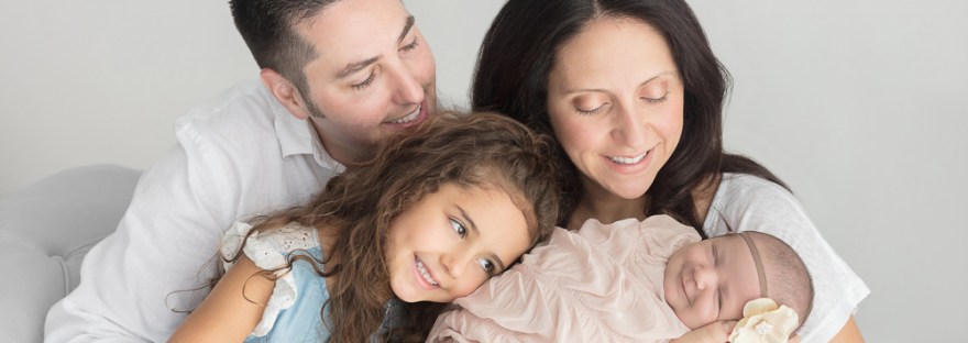 Smiling mommy, daddy and big sister posed with baby girl with white flower headband swaddled in champagne wrap for newborn portrait session