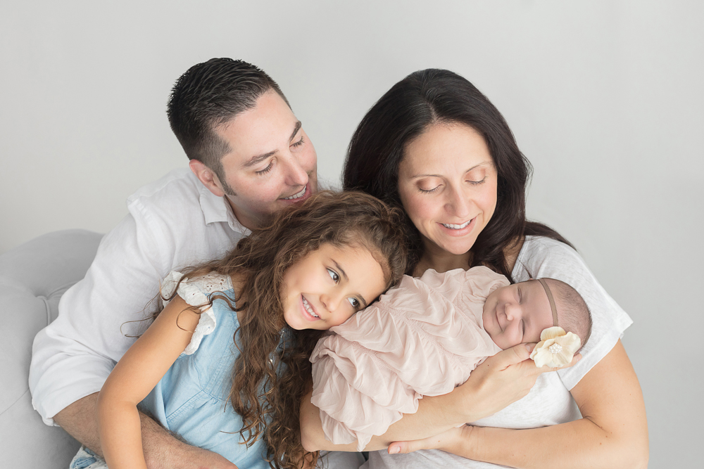 Smiling mommy, daddy and big sister posed with baby girl with white flower headband swaddled in champagne wrap for newborn portrait session