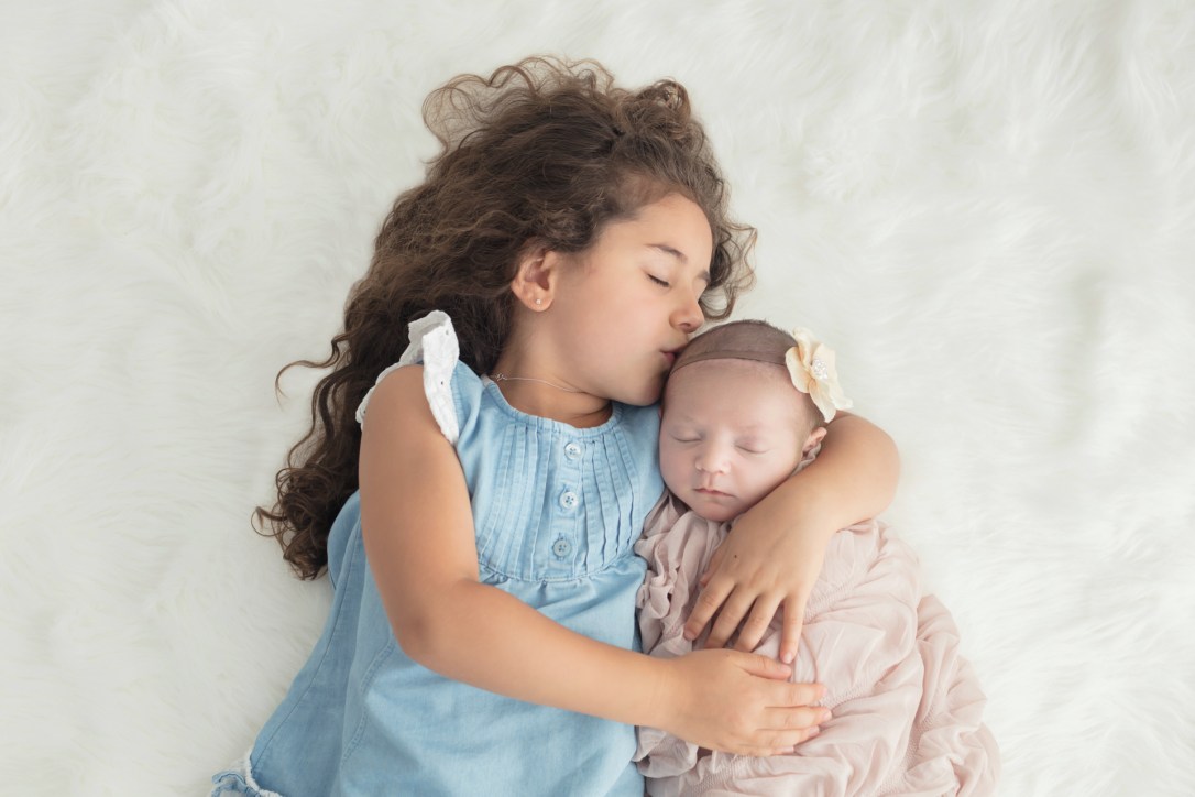 Big sister kissing new little sister with white flower headband swaddled in champagne wrap for baby girl's newborn portrait session