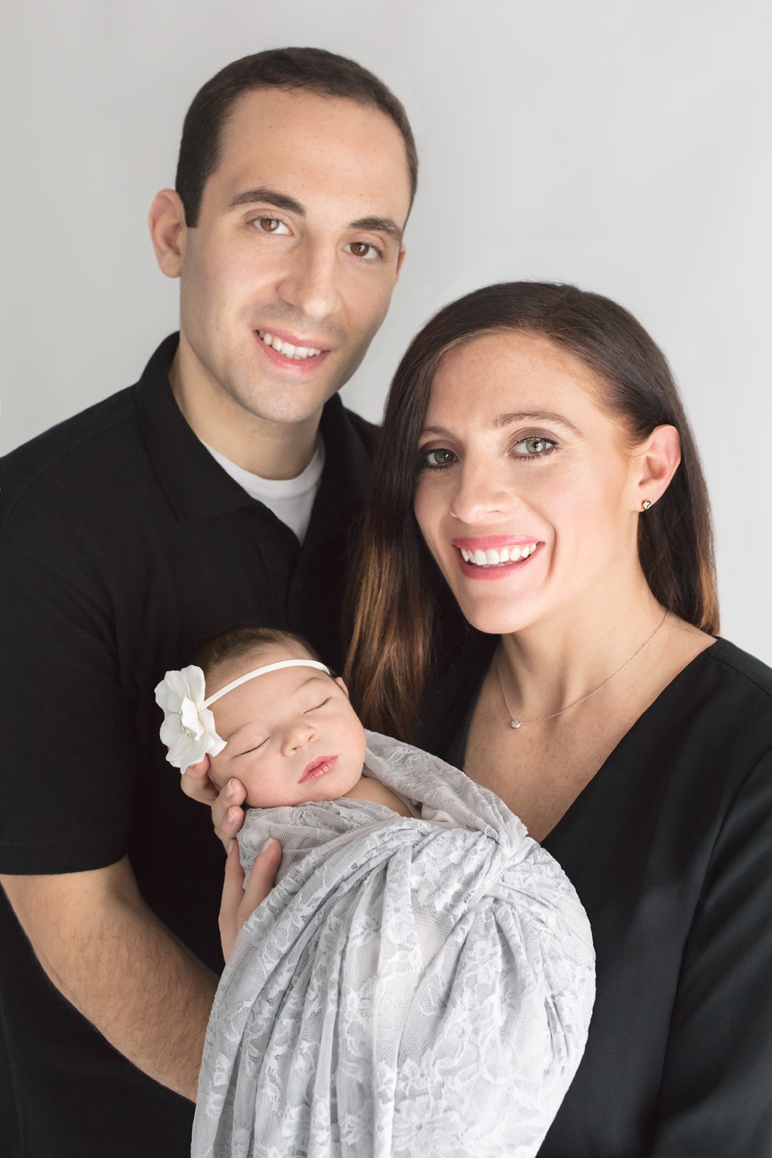 Happy mom and dad with first child, a baby girl, snuggled in lovely gray lace and white flower headband for newborn portrait session