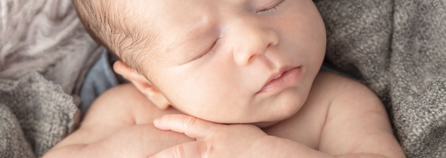 close image a newborn baby boy with hands clasped under his cheeks, wrapped in an open knit textured wool gray and denim swaddle for his newborn photography portrait at a Greenwich Connecticut newborn photography studio