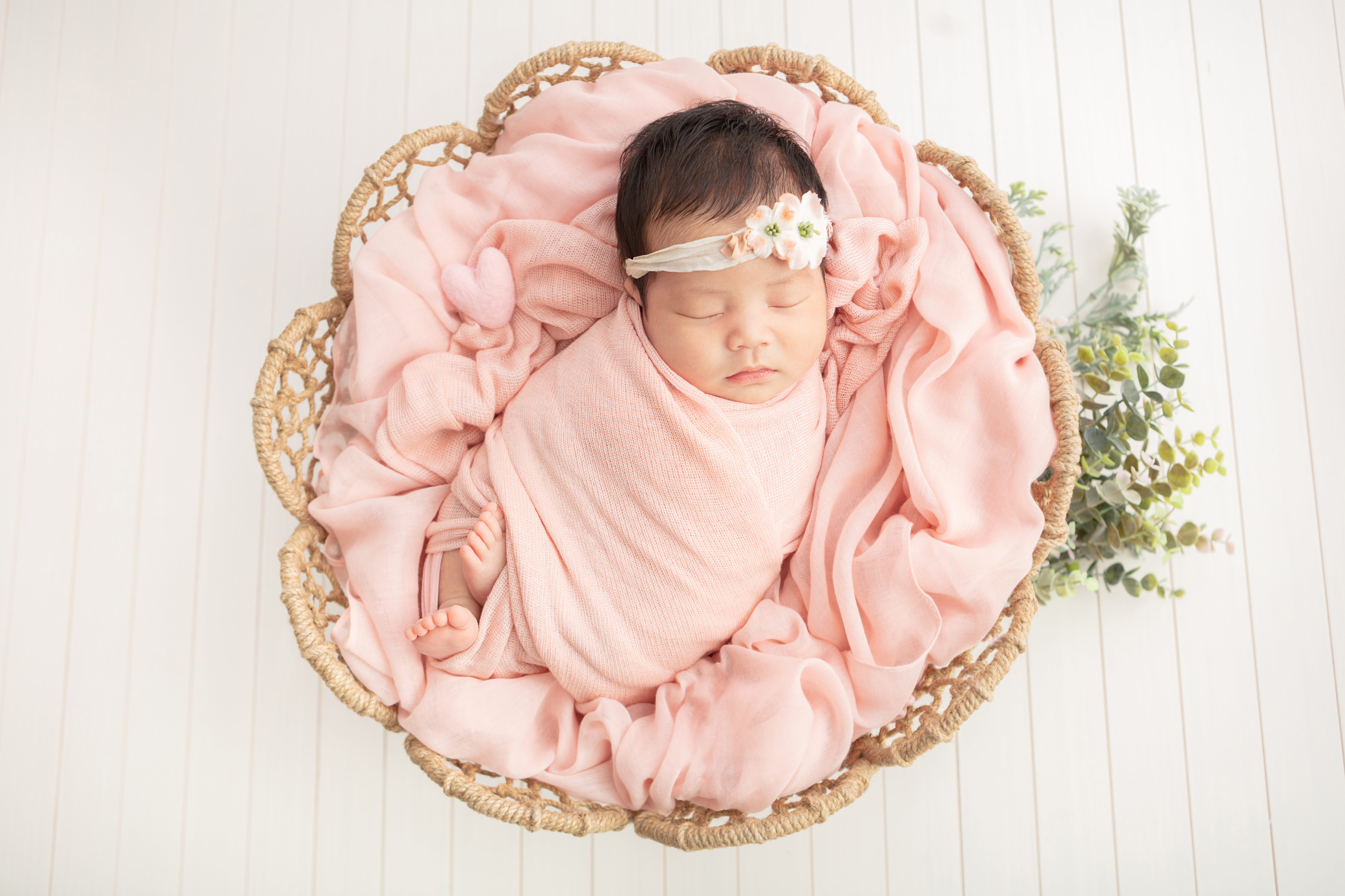 newborn baby girl with dark hair, swaddled in a light peachy pink swaddle, in a flower shaped wicker basket