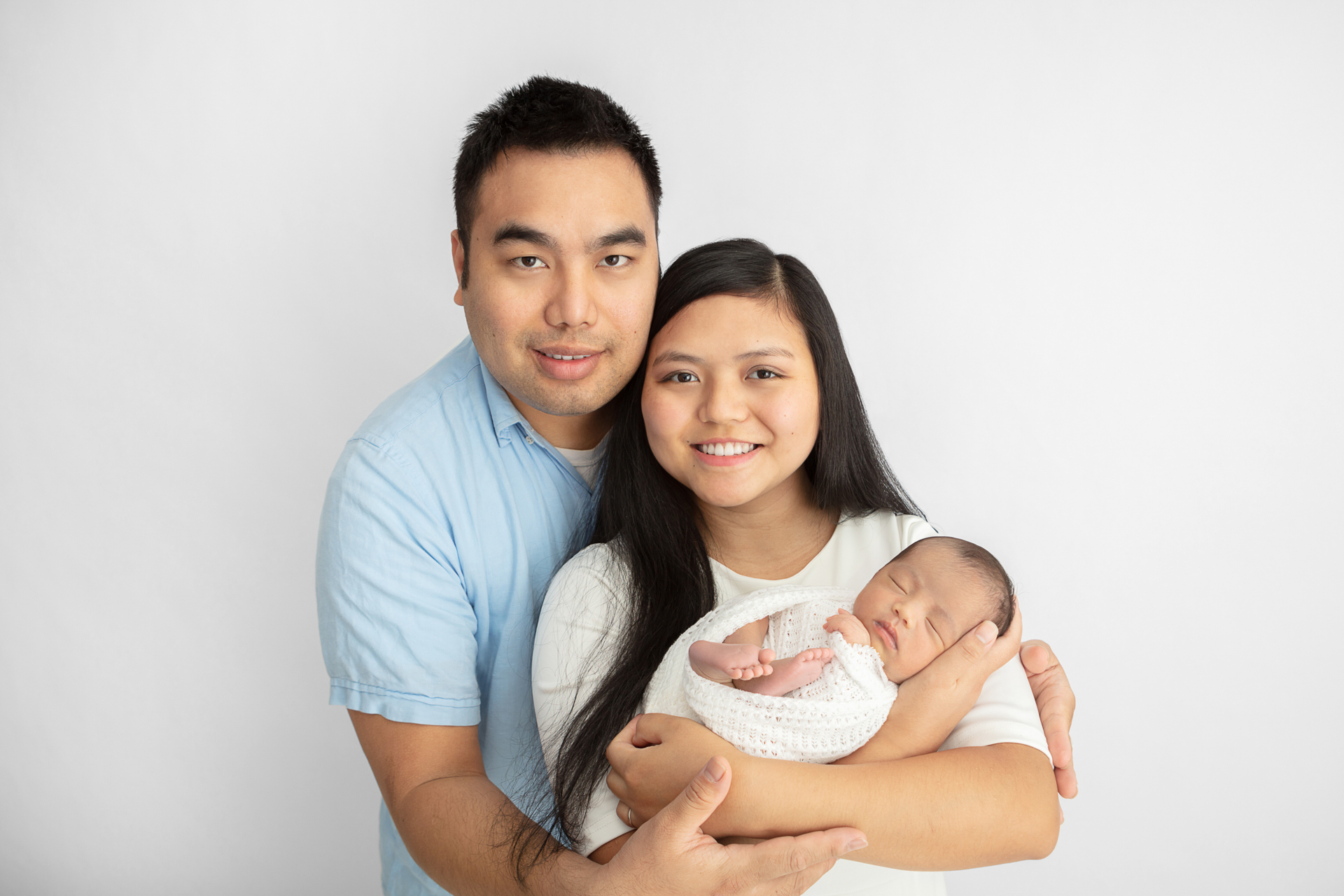 new family of three; new parents smiling at the camera; mom holding her newborn daughter as dad wraps his arms around his girls