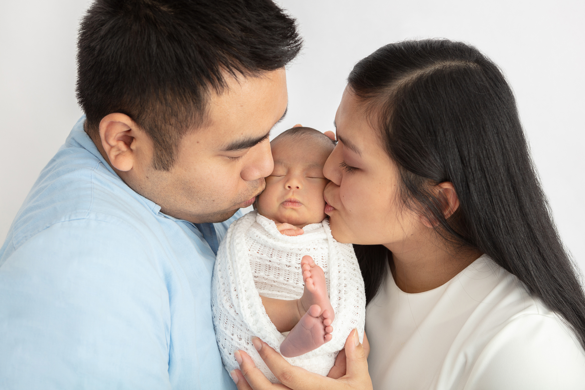 new father and mother kissing their newborn daughter's cheeks and holding her in between them
