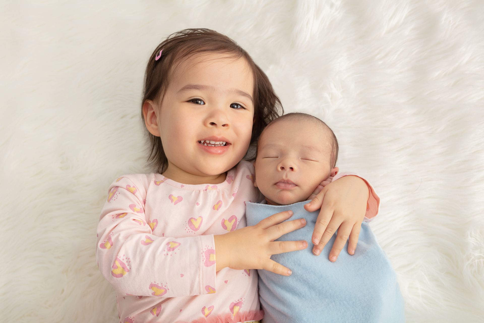 proud big sister wearing a pink long sleeved dress with pink and yellow hearts, with her arm around her baby brother