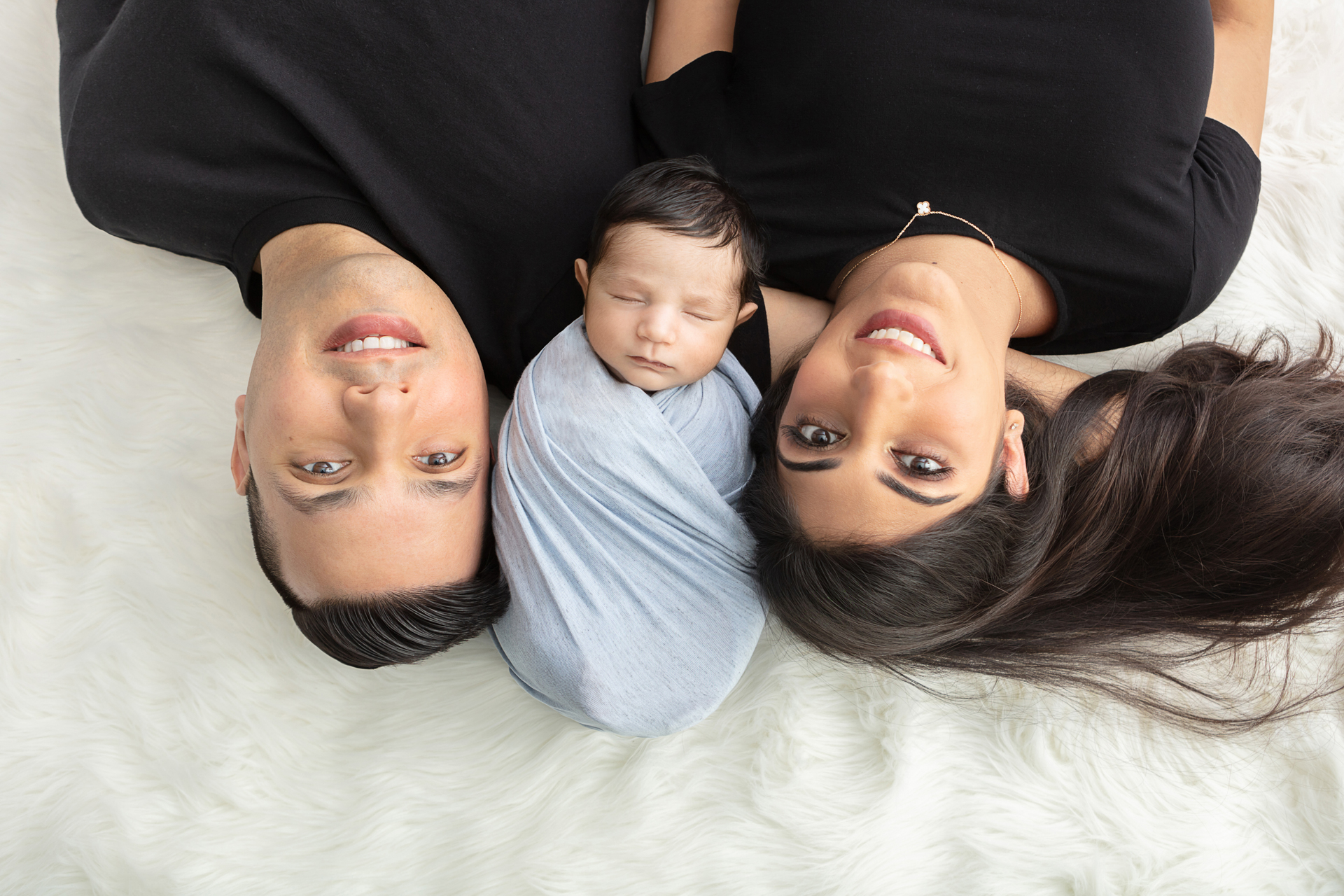 mom and dad wearing black tees lying down on a white flokati with their newborn in between them