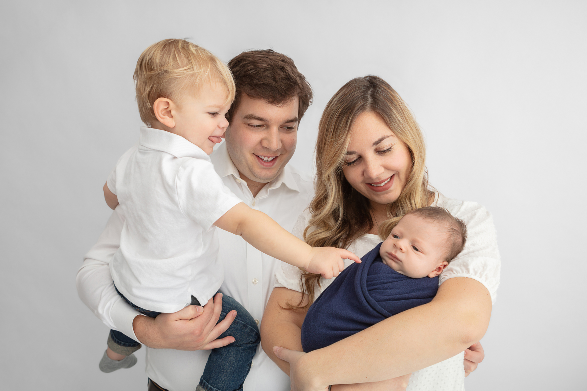 new family of four with toddler big brother sticking out his tongue and reaching out to touch his new baby brother