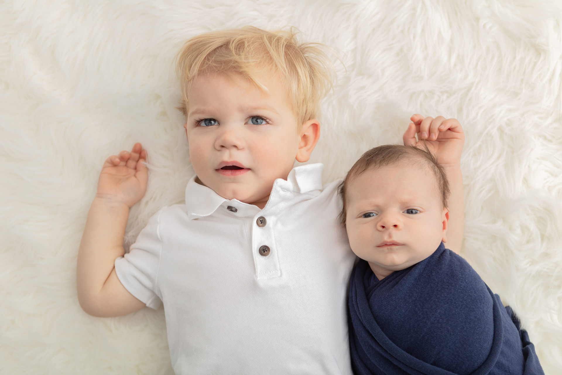 newborn baby boy with a concerned look on his face, wrapped in a navy swaddle; big brother with blonde hair with his new baby brother