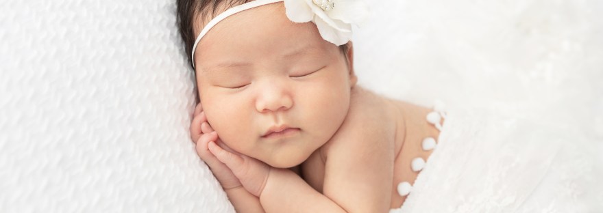 newborn baby girl with dark hair wrapped in a white lace fabric cradled on a soft white fabric withy a white flower headband, hands clasped under her cheek in a sleepy pose