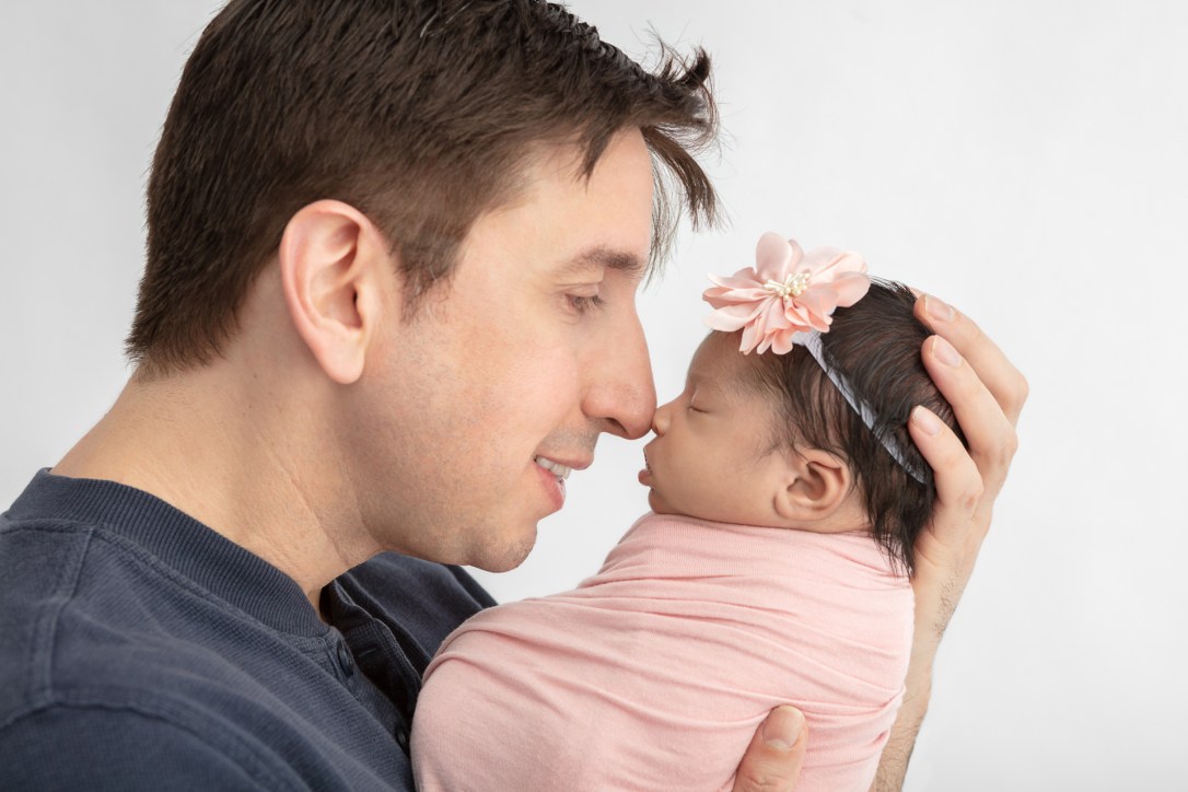 new dad touching noses with his newborn baby girl who is swaddled in pink and wearing a large pink flower headband