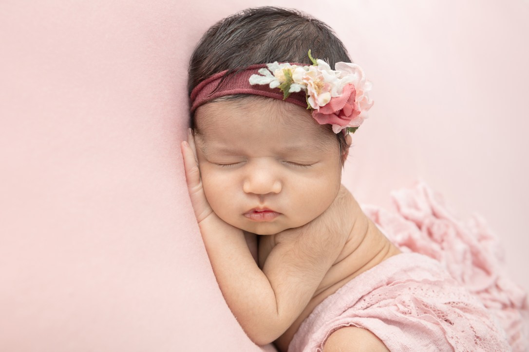 newborn baby girl with a lot of dark hair asleep against a pink pillow and wearing a pink floral headband