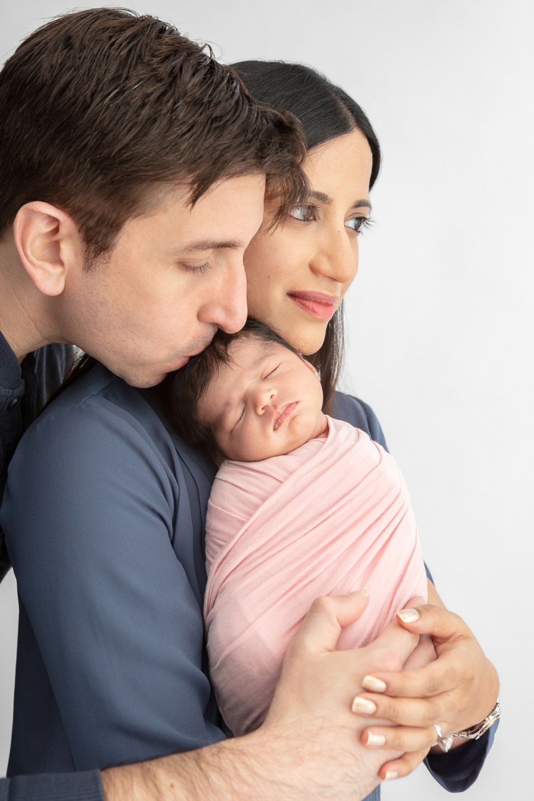 new mom with big dark eyes looking into the distance as she holds her newborn daughter while dad kisses the baby's head