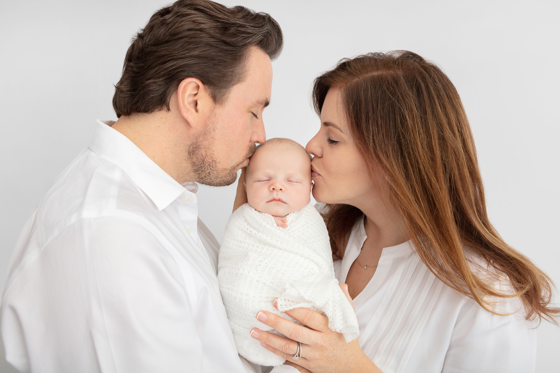 new father and mother dressed in white kissing their newborn baby boy who is being held between them