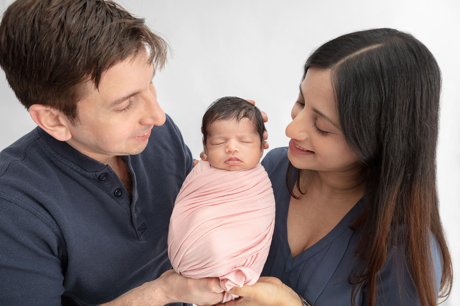 new mom and dad wearing dusty blue shirts holding up their newborn baby girl in between them